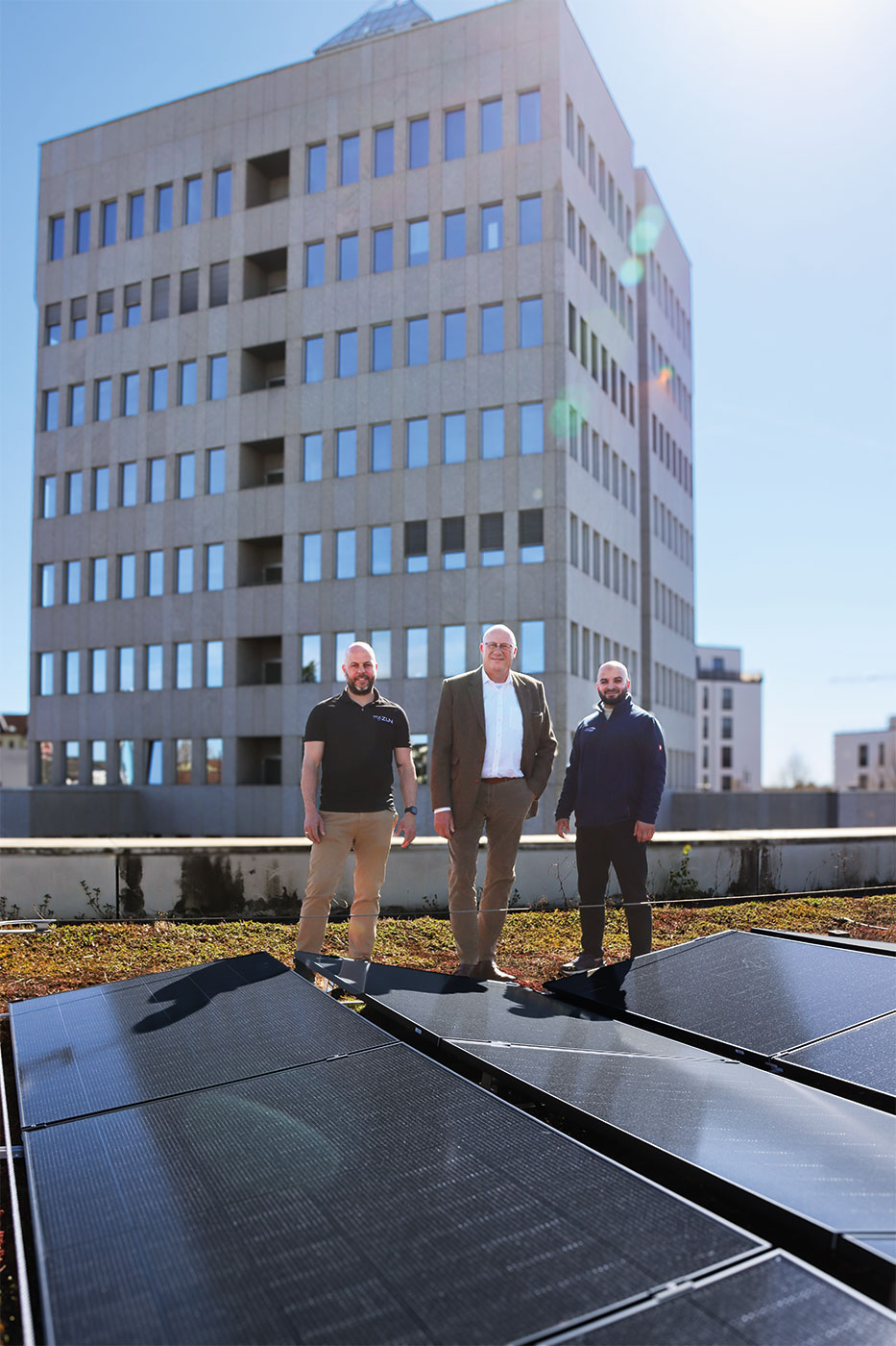 3 Männer stehen hinter ein paar PV-Modulen auf dem Flachdach eines Gebäudes.