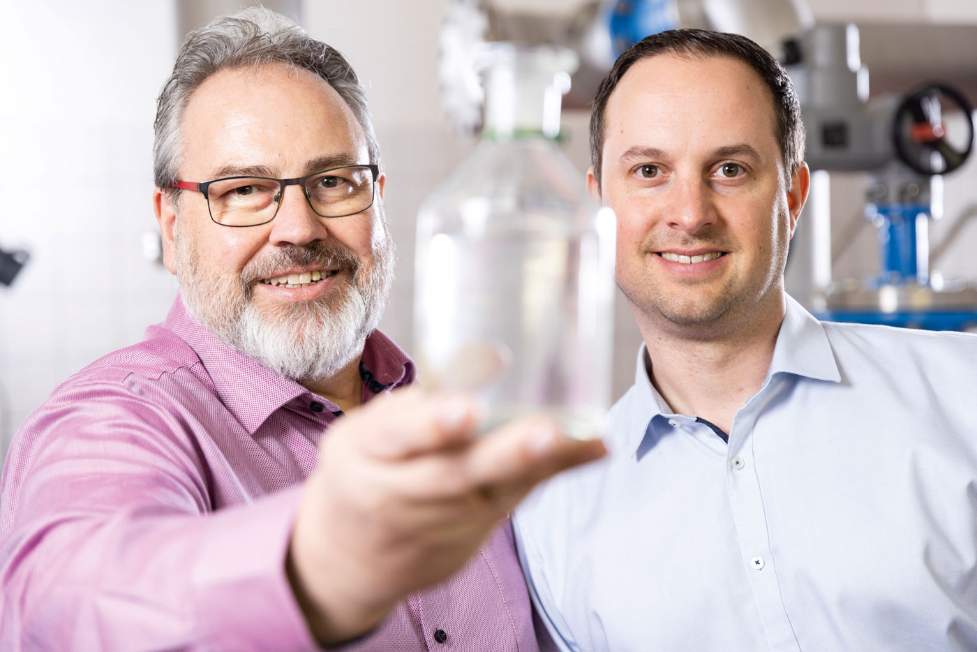 Zwei Männer stehen nebeneinander und schauen in die Kamera. Einer hält eine Glasflasche mit Wasser nah an die Kamera