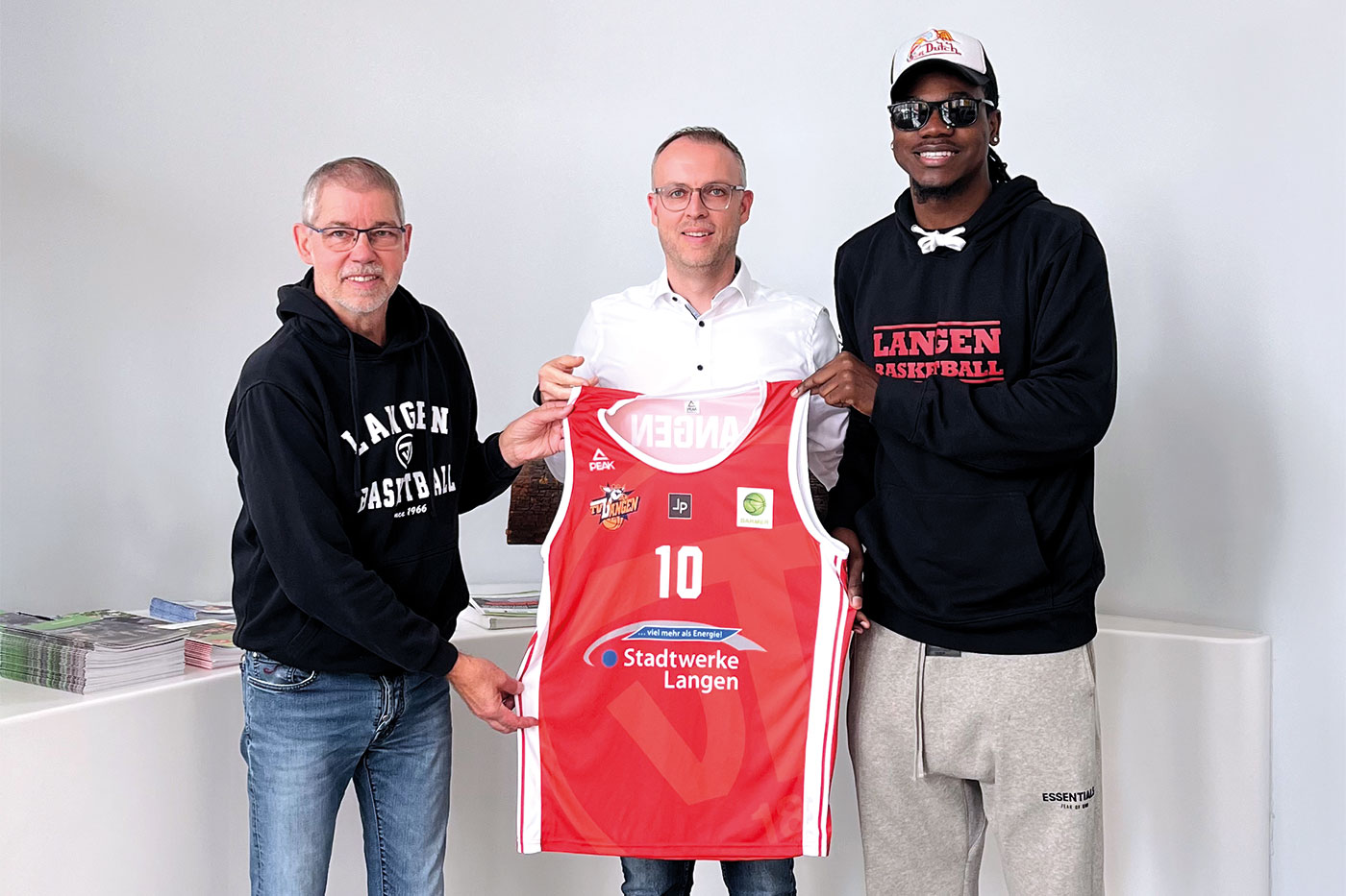 Drei Männer stehen nebeneinander und halten ein Basketball-Trikot hoch.