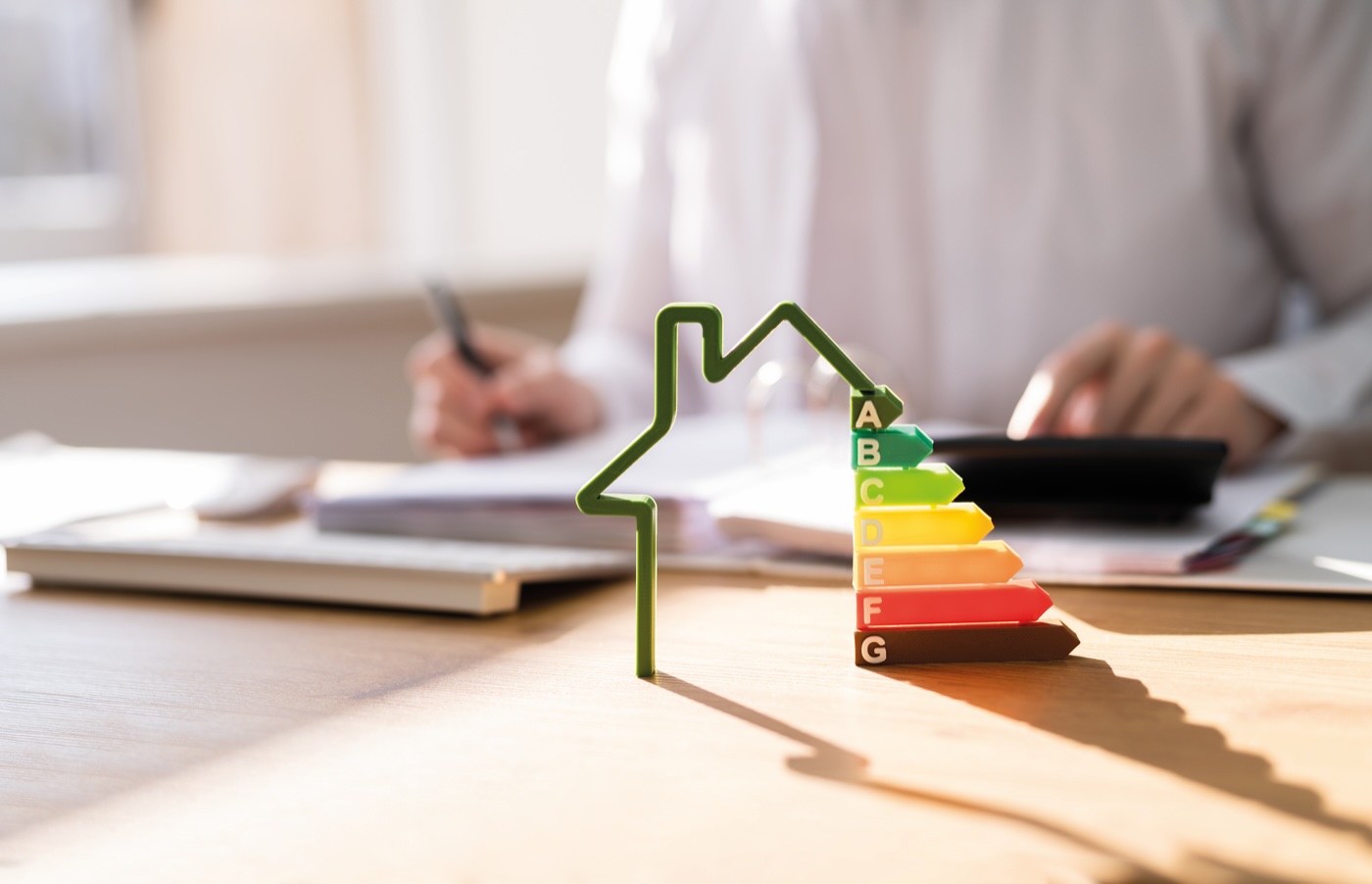 Verschwommen im Hintergrund: Eine Person arbeitet an einem Schreibtisch. Im Vordergrund eine Haus-Grafik kombiniert mit den Energieeffizienz-Klassen A-G