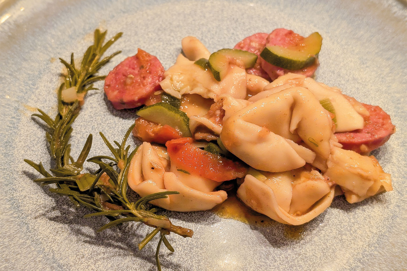 Mediterraner Tortellini-Eintopf auf einem blauen Teller angerichtet und einem Rosmarin-Zweig.
