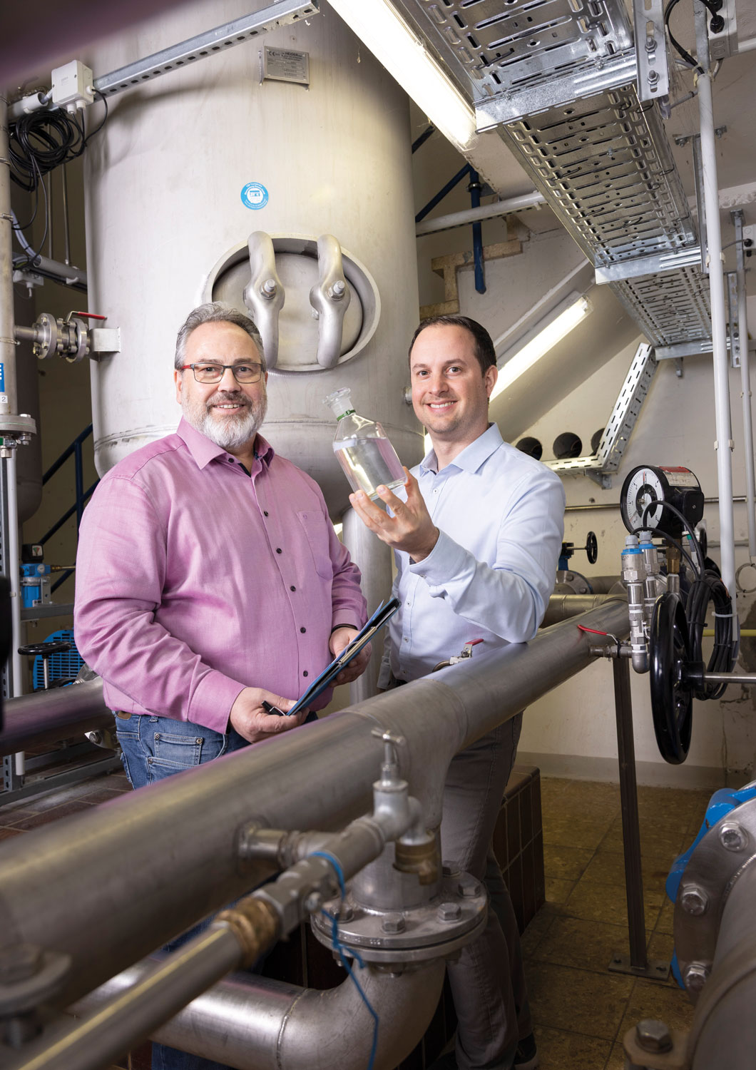 zwei Männder stehen vor einem großen Trinkwasserbehälter im Wasserwerk. Einer hält eine Glasflasche mit Wasser hoch.