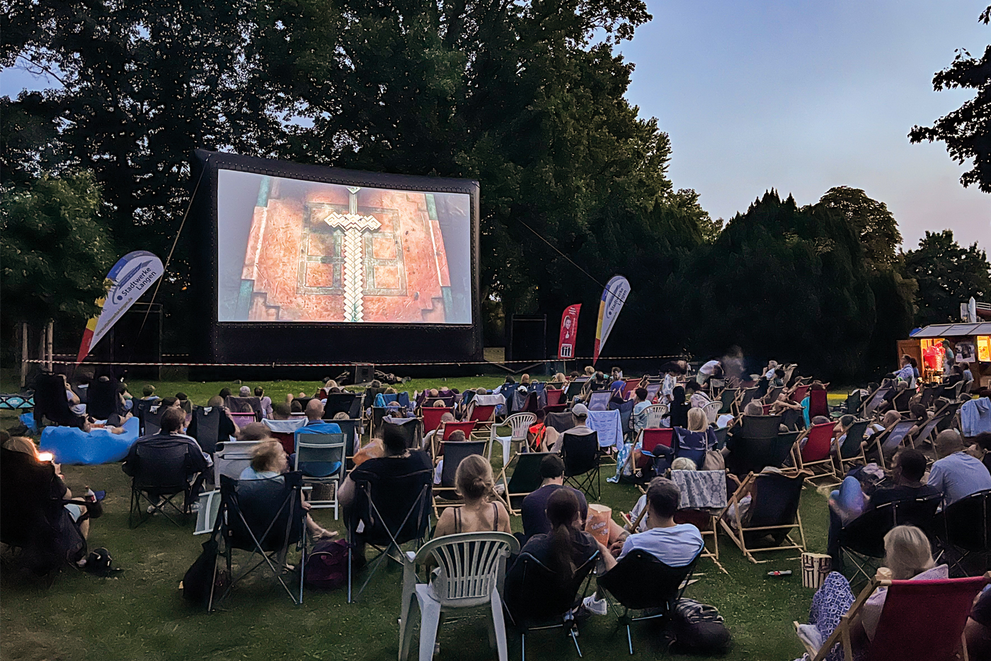 Viele Menschen sitzen auf Stühlen und Decken im Freien und schauen bei Dämmerung einen Film auf einer großen Leinwand in einem Freibad.