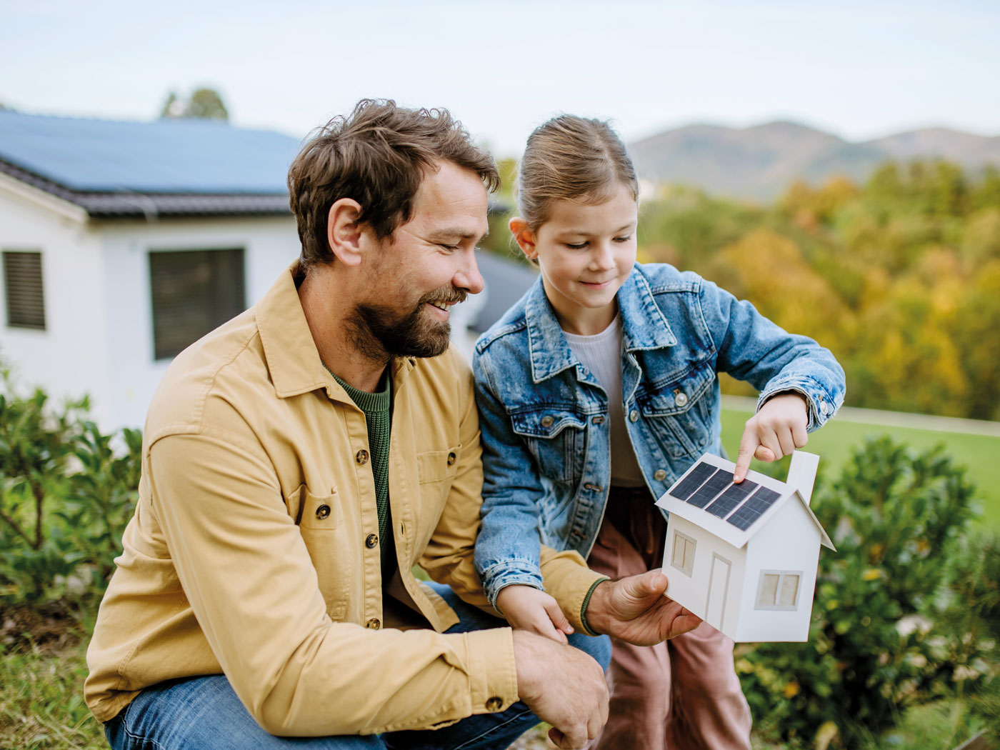 Mann und Kind sitzen im Garten und halten ein Papierhaus mit PV-Anlage in der Hand.