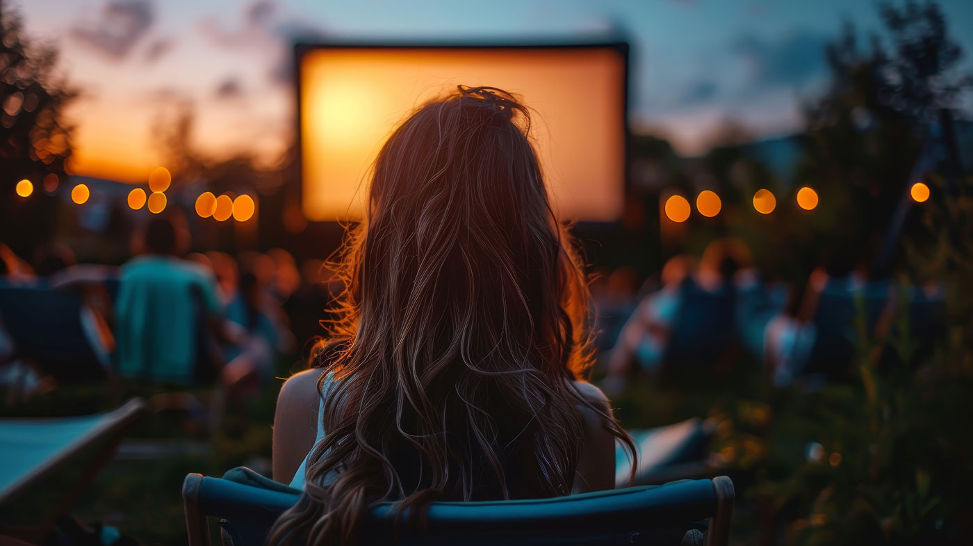 Open Air Kino in einer klaren Sommernacht. Sicht auf eine Frau mit langen Haaren von hinten. Sie schaut auf eine Leinwand.