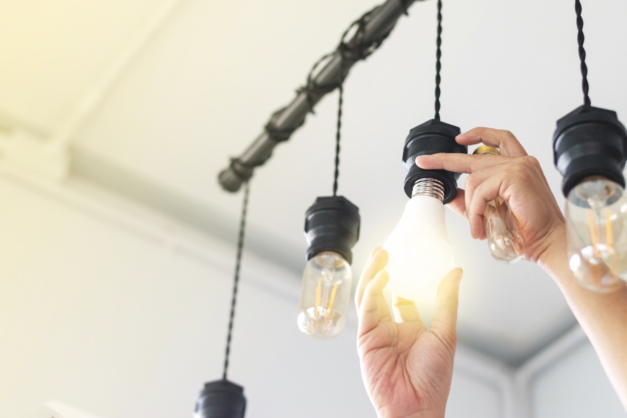Eine Hand dreht eine neue Glühbirne in die Halterung einer hängenden Deckenlampe. Die neu eingedrehte Glühbirne Leuchtet, die anderen sind aus.