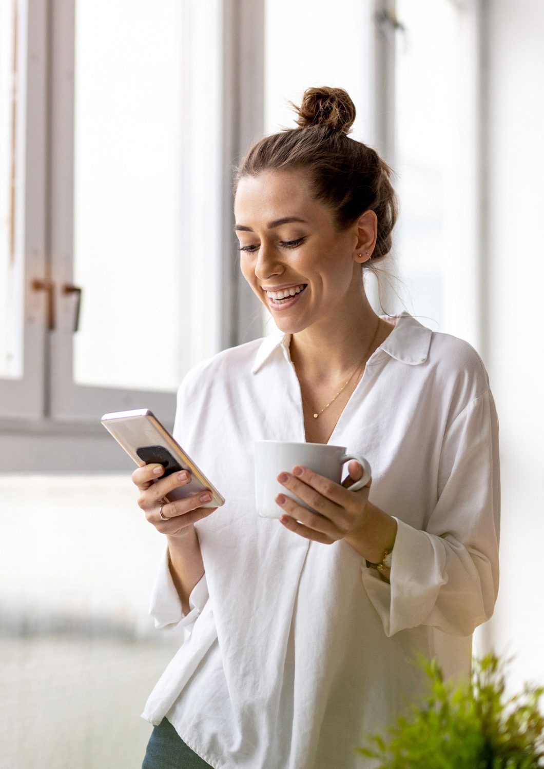 Junge Frau mit weißer Bluse steht mit einem Handy in der einen Hand und einer Tasse in der anderen vor großen Fenstern und Tippt am Handy.