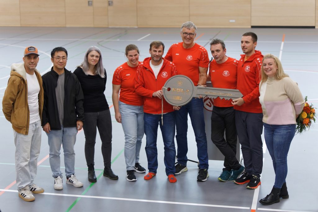 Vier-Felder-Halle eingeweiht 2 Eine Gruppe an Menschen stehen in einer Turnhalle und halten einen großen symbolischen Schlüssel aus Holz in den Händen.