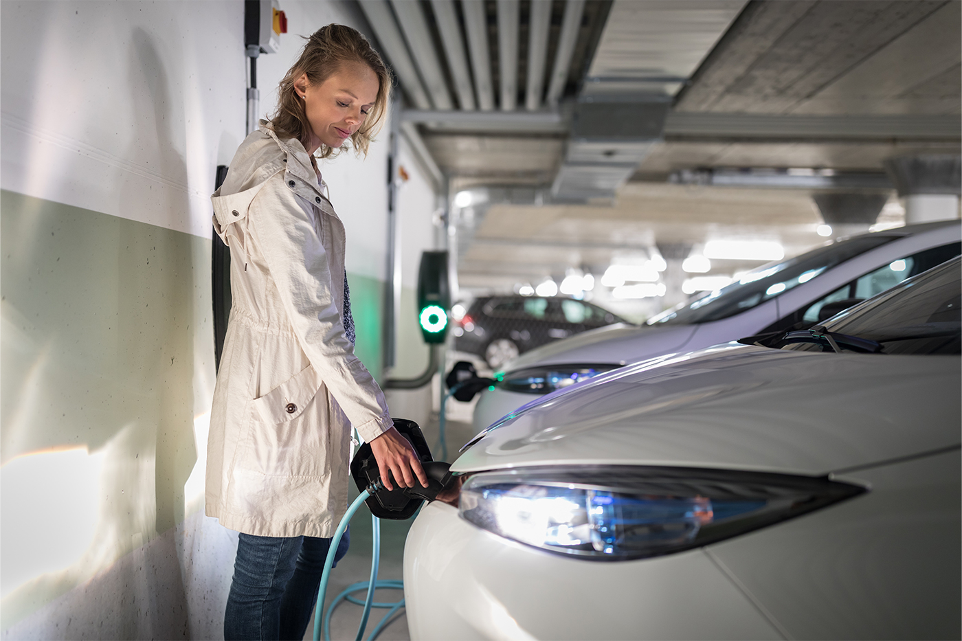 Eine junge Frau mit einem hellen Mantel lädt ein Elektroauto in einer Tiefgarage auf.