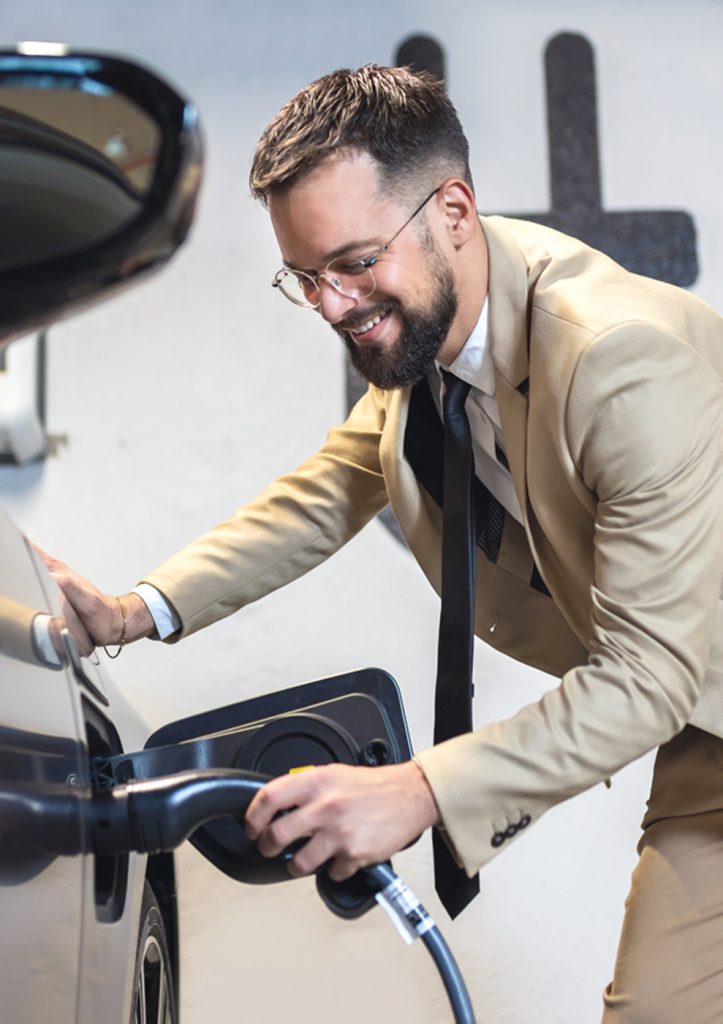 Ein junger Mann in einem hellen Anzug steckt ein Ladekabel in ein Elektroauto.