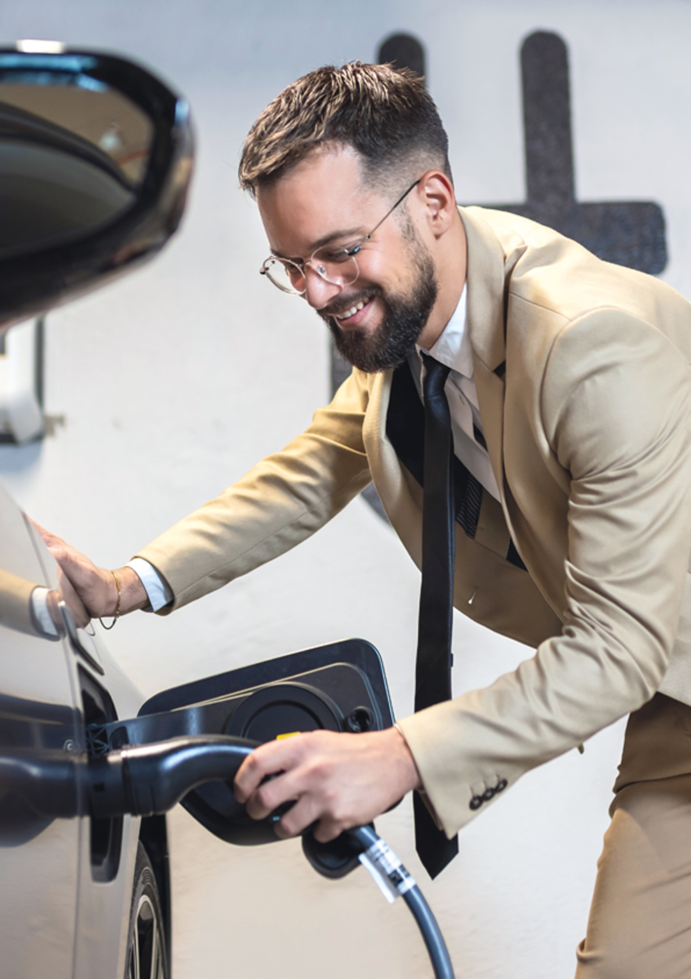 Ein junger Mann in einem hellen Anzug steckt ein Ladekabel in ein Elektroauto.