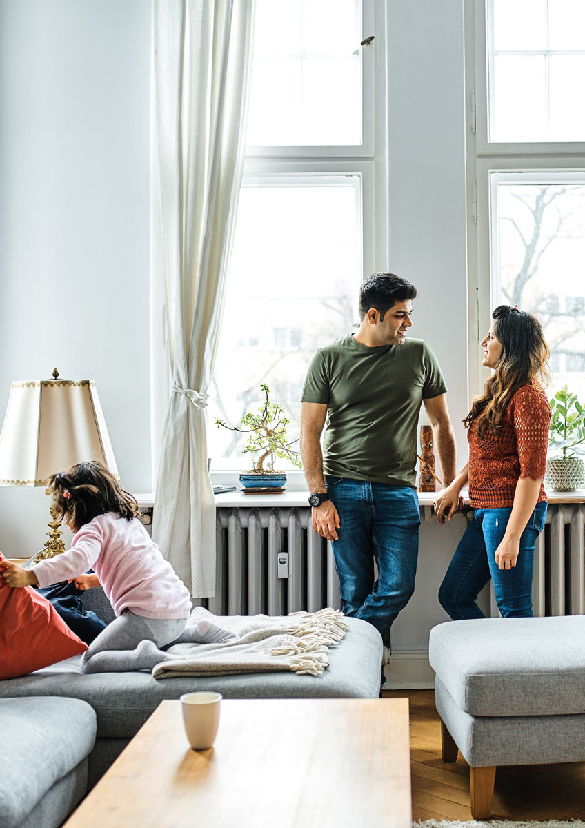 Mann und Frau stehen an einem Fenster in einer Altbauwohnung und schauen sich an. Im Vordergrund spielt ein Kind auf dem Sofa.